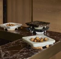 Three Caldera marble bowls by Greg Natale, each square in shape and evocative of rationalist architecture, display small brown fruits atop a dark marble countertop within a modern space featuring wooden walls and stacked marble trays in the background.