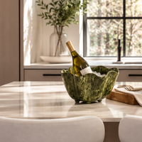 A bottle of white wine chills in the Beatriz Ball Resin Malachite Ice Bucket - Green, filled with ice and placed on a marble kitchen island. In the background, a vase of green branches sits near a sunlit window, while two white chairs are visible in the foreground.