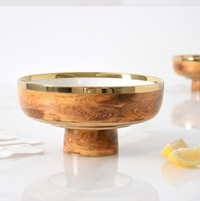 The Madera Footed Bowl - by PAMPA BAY features a wooden base, gold-toned rim, and white interior, styled with a lemon wedge and napkin on a white surface; another bowl is blurred in the background.