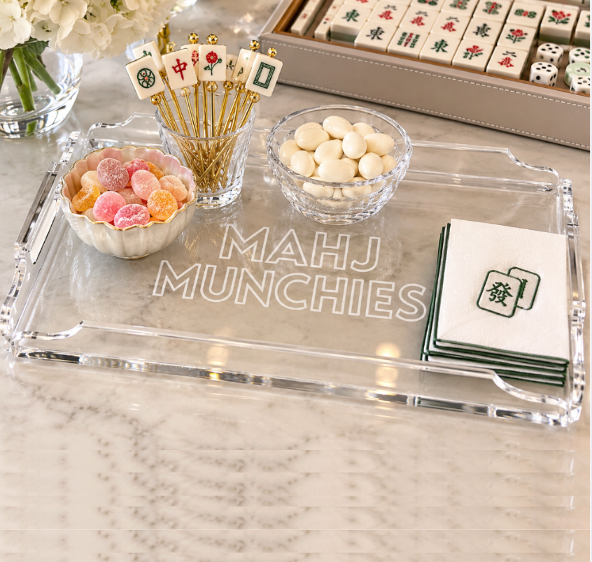 The Mahj Munchies Game Tray by Eye Candy displays candy, white chocolates, cocktail picks, and Mahjong-themed napkins on marble, accented with Mahjong tiles and flowers.
