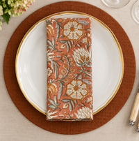 A folded rust-colored Block Print Napkin Set of 4 - Rust with a floral block print pattern resting on a white dinner plate with a gold rim.