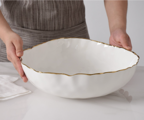 A person in a striped apron holds the PAMPA BAY Portofino Serving Bowl with elegant gold trim on a white marble surface. A folded white cloth nearby highlights the beauty of this oven to table collection piece.