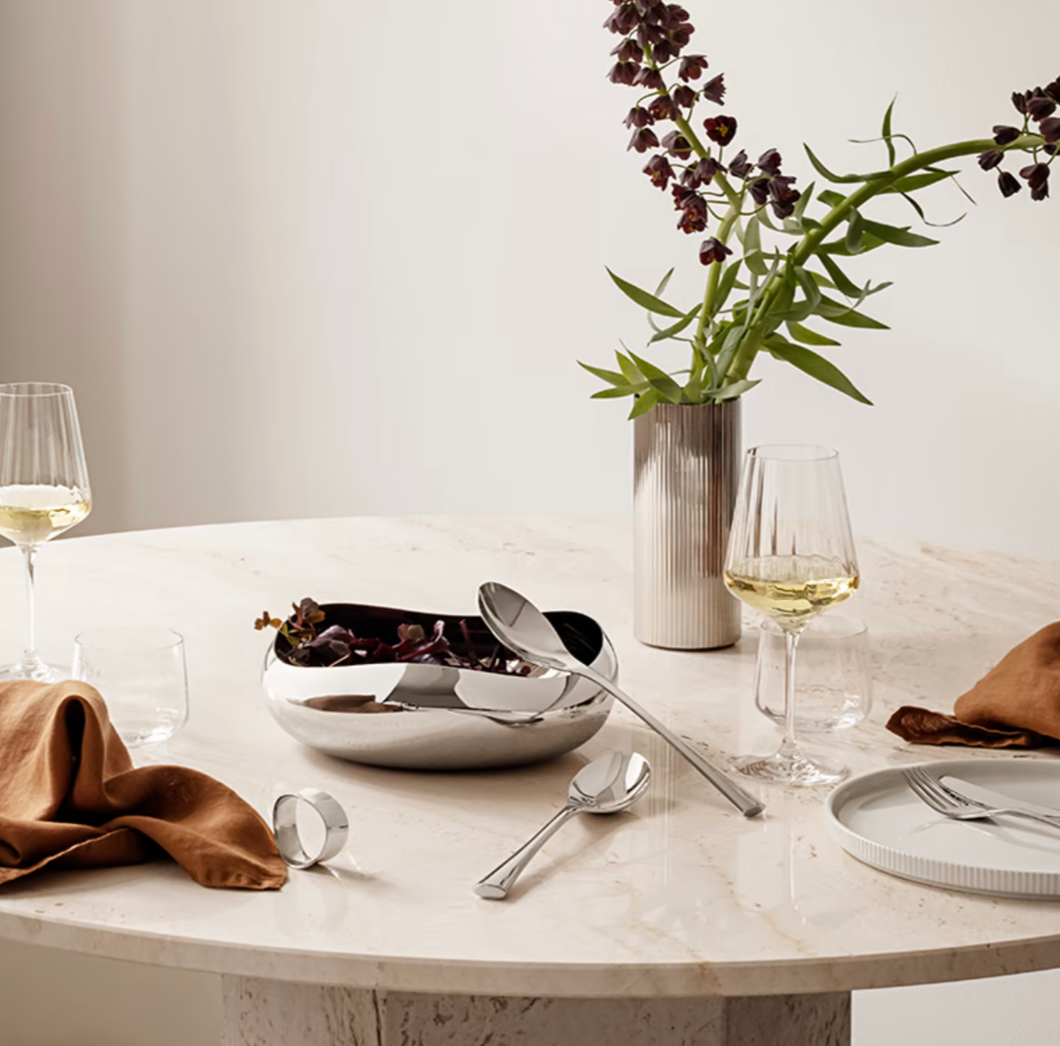 A round marble table set with wine glasses, silver utensils, a napkin, the Georg Jensen Cobra Salad Server Set including a serving fork and spoon, and a silver bowl with dark flowers beside a tall vase holding purple flowers against a light background.
