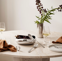 A round marble table set with wine glasses, silver utensils, a napkin, the Georg Jensen Cobra Salad Server Set including a serving fork and spoon, and a silver bowl with dark flowers beside a tall vase holding purple flowers against a light background.