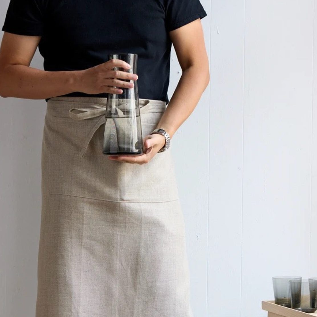 A person in a beige apron and black shirt holds a hand blown glass carafe with dark liquid. Their torso and arms are visible. Two Gary Bodker Everyday Glasses Charcoal rest on a wooden surface in the lower right corner.