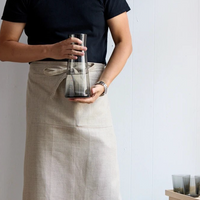 A person in a beige apron and black shirt holds a hand blown glass carafe with dark liquid. Their torso and arms are visible. Two Gary Bodker Everyday Glasses Charcoal rest on a wooden surface in the lower right corner.