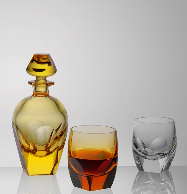 The Moser Bar Decanter 750 ml in yellow with a matching stopper stands beside two short glasses—one filled with amber liquid, the other empty—against a plain light gray background.