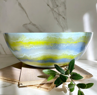 The Redondo Bowl Portofino Blue by CDMX / PAOLA VALLE, crafted from food safe resin, features wavy blue, yellow, and white patterns. It sits on wooden tiles beside a green branch and marble background in gentle sunlight.