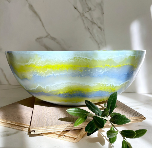 The Redondo Bowl Portofino Blue by CDMX / PAOLA VALLE, crafted from food safe resin, features wavy blue, yellow, and white patterns. It sits on wooden tiles beside a green branch and marble background in gentle sunlight.