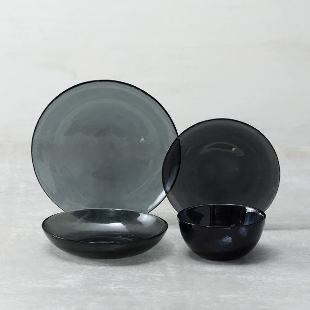 The Fortessa Los Cabos Shadow Dinnerware 4 Piece set features translucent black and gray hammered glass—plate, salad plate, shallow bowl, and small bowl—arranged on a light gray surface against a pale background.