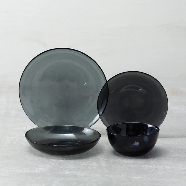 The Fortessa Los Cabos Shadow Dinnerware 4 Piece set features translucent black and gray hammered glass—plate, salad plate, shallow bowl, and small bowl—arranged on a light gray surface against a pale background.