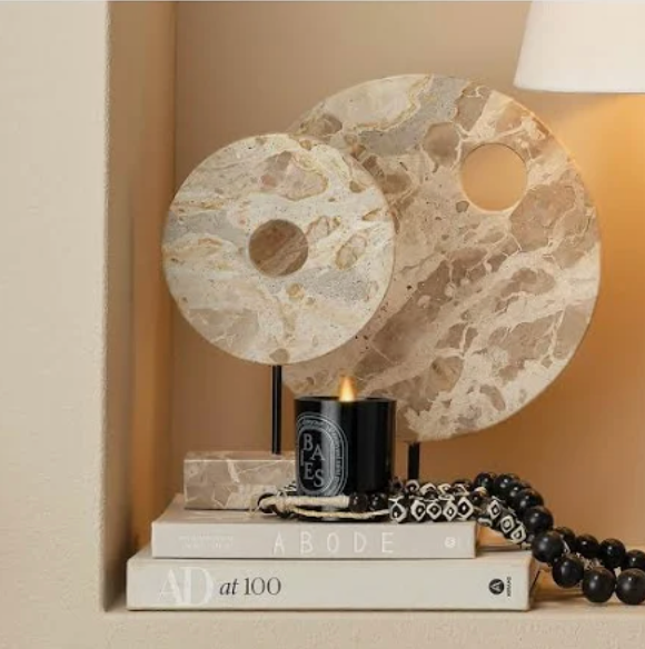 The Marble Cream Circle Disk on Stand by Jamie Young is showcased above stacked books, next to a lit black candle and black beads, with a lamp nearby, creating a celestial-inspired display.
