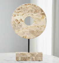 The Marble Cream Circle Disk on Stand by Jamie Young features a round, cream marble sculpture with a center hole, mounted on a black rod and rectangular stone base—ideal for adding celestial decor to your space.