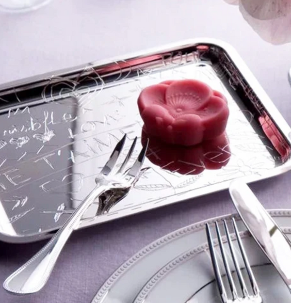 A pink, flower-shaped dessert sits on the LOVE Graffiti Christofle Silver Plated Tray by Christofle, paired with a dessert fork. The shiny tray on a lavender tablecloth adds playful elegance to the scene.