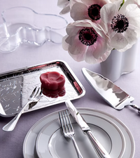 A delicate pink flower-shaped confection rests on a Christofle LOVE Graffiti Silver Plated Tray beside white anemones. The elegant scene is completed with formal silverware, plates, and a cake server.