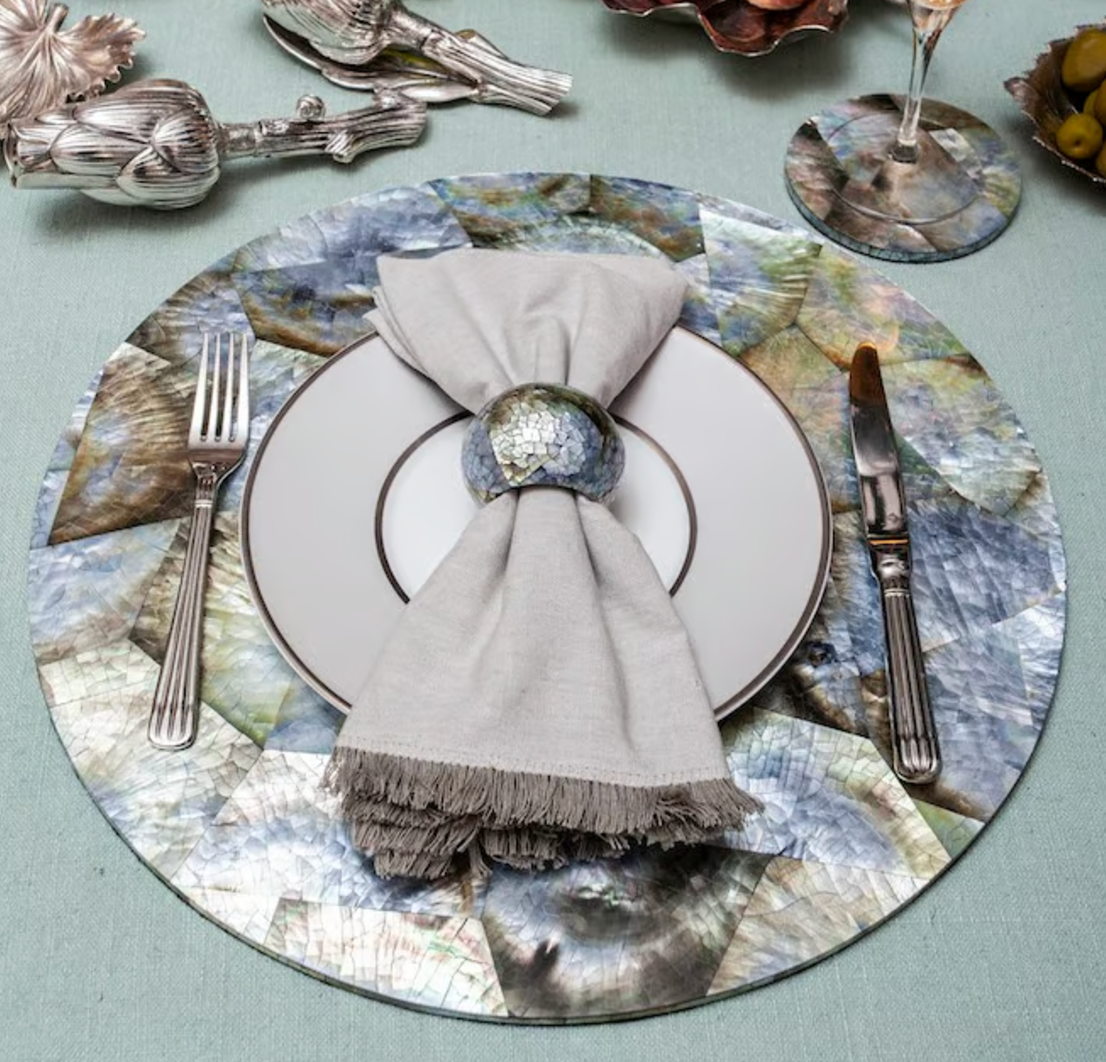 A decorative table setting featuring a silver fork and knife, a white plate, and a beige napkin accented with the Von Gern Home Shell Pearl Black Napkin Ring (Set of 4), all arranged on a colorful iridescent placemat atop a light blue tablecloth.