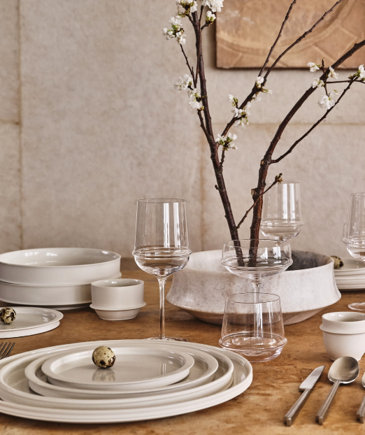 A table set with white dishes, Dune White Wine Glasses by Serax, cutlery, and a centerpiece of blossom branches in a vase. Quail eggs decorate the plates. The setting has a minimalist, elegant style.