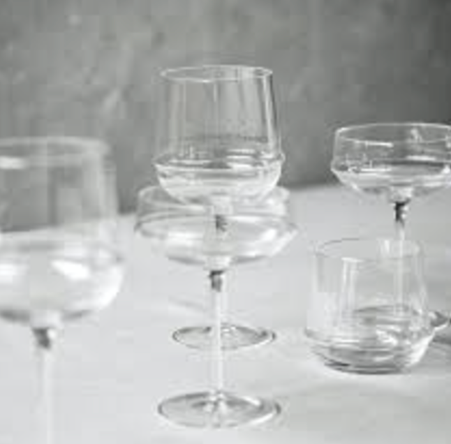 A collection of clear, empty glassware—including the Serax Dune White Wine Glass, coupe glasses, and a tumbler—arranged on a white surface against a softly blurred gray background.