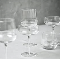 A collection of clear, empty glassware—including the Serax Dune White Wine Glass, coupe glasses, and a tumbler—arranged on a white surface against a softly blurred gray background.