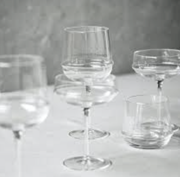 A collection of clear, empty glassware—including the Serax Dune White Wine Glass, coupe glasses, and a tumbler—arranged on a white surface against a softly blurred gray background.