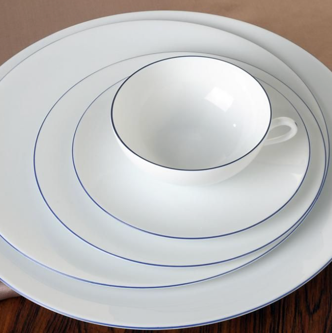 A white teacup with a handle sits on a saucer, stacked on two larger Raynaud Monceau Bleu Outremer porcelain plates featuring thin ultramarine blue rims, all arranged on a wooden table.