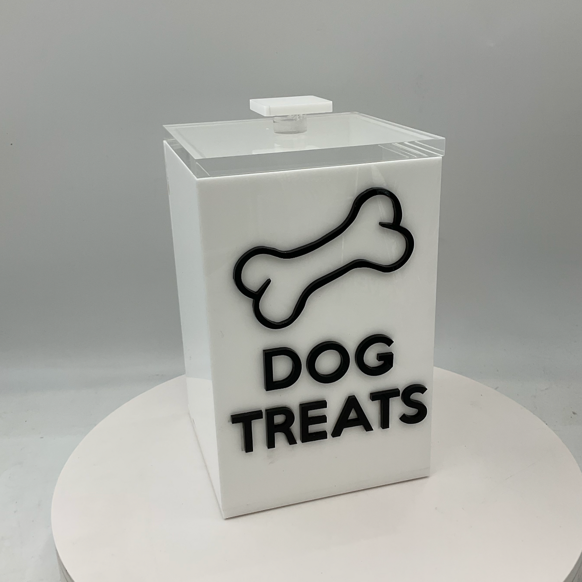 The Eye Candy Dog Treats Canister is a white rectangular holder with a clear lid, labeled "DOG TREATS" in bold black letters and decorated with a black dog bone outline.