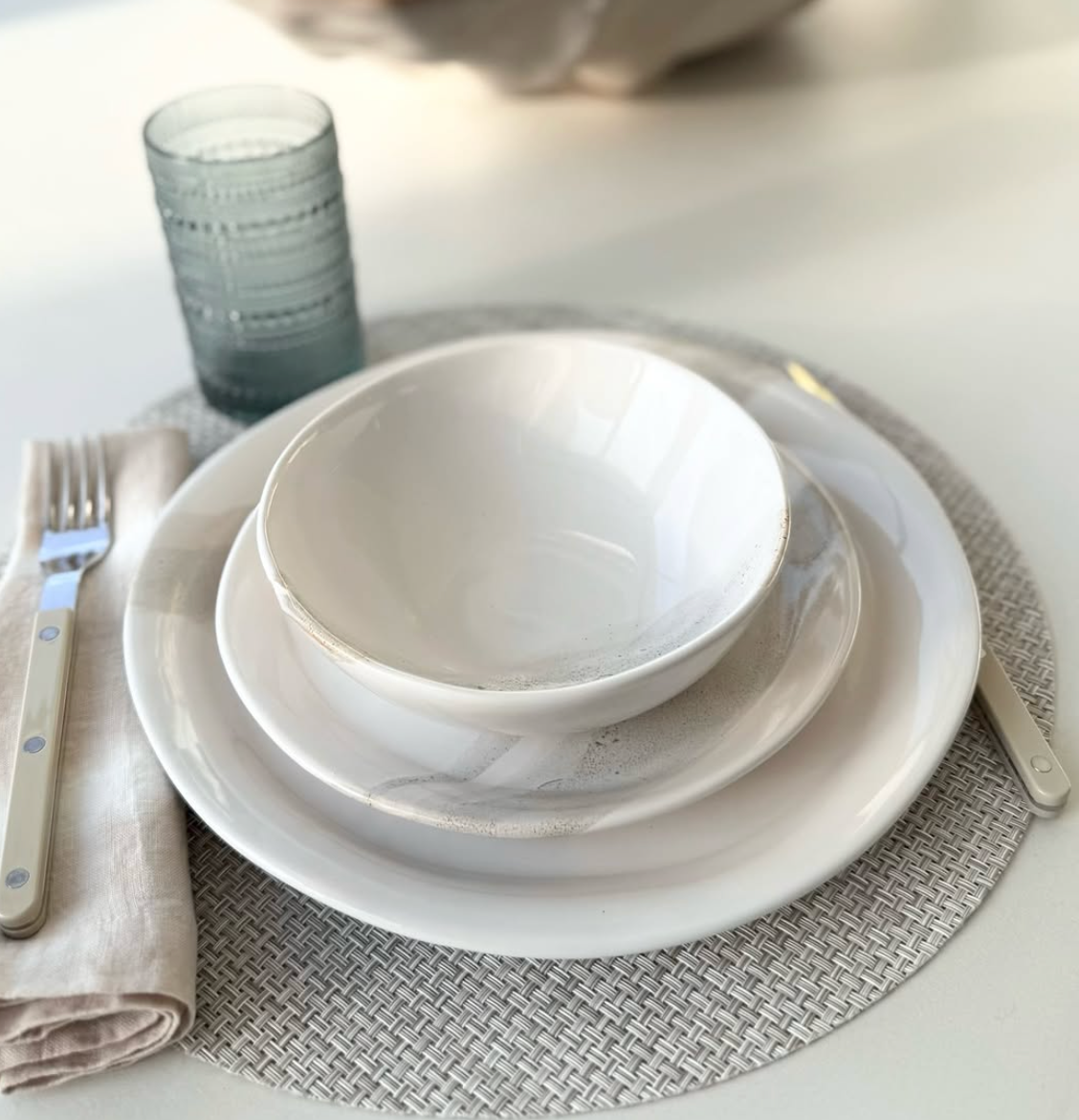 A neatly set table features the Urban Dinnerware - Custom by Alex Marshall Studios, including handmade stoneware plates, a salad plate, bowl, beige napkin with cutlery, a round placemat, and a textured glass tumbler on a white surface.