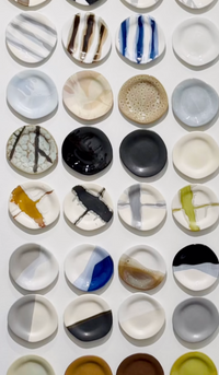 A grid of Urban Dinnerware - Custom plates by Alex Marshall Studios, each handmade with unique abstract patterns and colors, is displayed on a white wall. These microwave and dishwasher safe plates feature organic stripes, splatters, and color blocks.