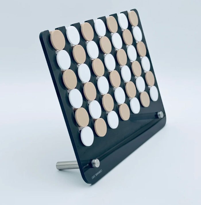 The Connect Four El Cuatro by Luxe Dominoes is a modern take on the classic four in a row game, featuring a black frame with alternating rows of white and beige circular pieces, elegantly displayed on a metal stand against a light background.