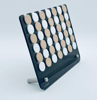 The Connect Four El Cuatro by Luxe Dominoes is a modern take on the classic four in a row game, featuring a black frame with alternating rows of white and beige circular pieces, elegantly displayed on a metal stand against a light background.