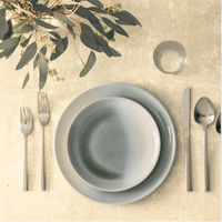 Overhead view of a minimalist table set with two Fortessa La Cotera Melamine Dinnerware Gray plates, flatware, a glass of water, and eucalyptus branches on a light surface.