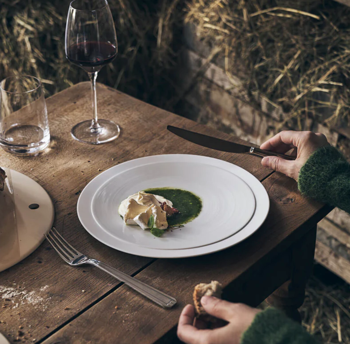 A person sits at a rustic wooden table set with Degrenne’s Normandy Flatware 5 Piece Set, savoring gourmet food with green sauce and a creamy topping, accompanied by red wine and hay visible in the background.