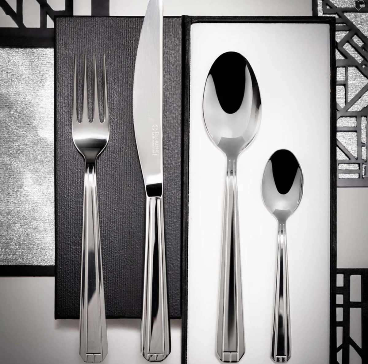 The Degrenne Normandy Flatware 5 Piece Set, featuring stylish Art Deco silverware, is displayed on a black and white geometric-patterned background.