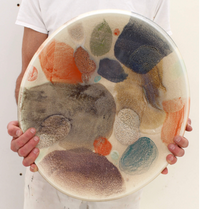 A person in a white shirt holds the KleinReid Rio Abrico Platter X Large, featuring abstract multicolored splotches and organic shapes in orange, blue, green, brown, and purple on a light background.