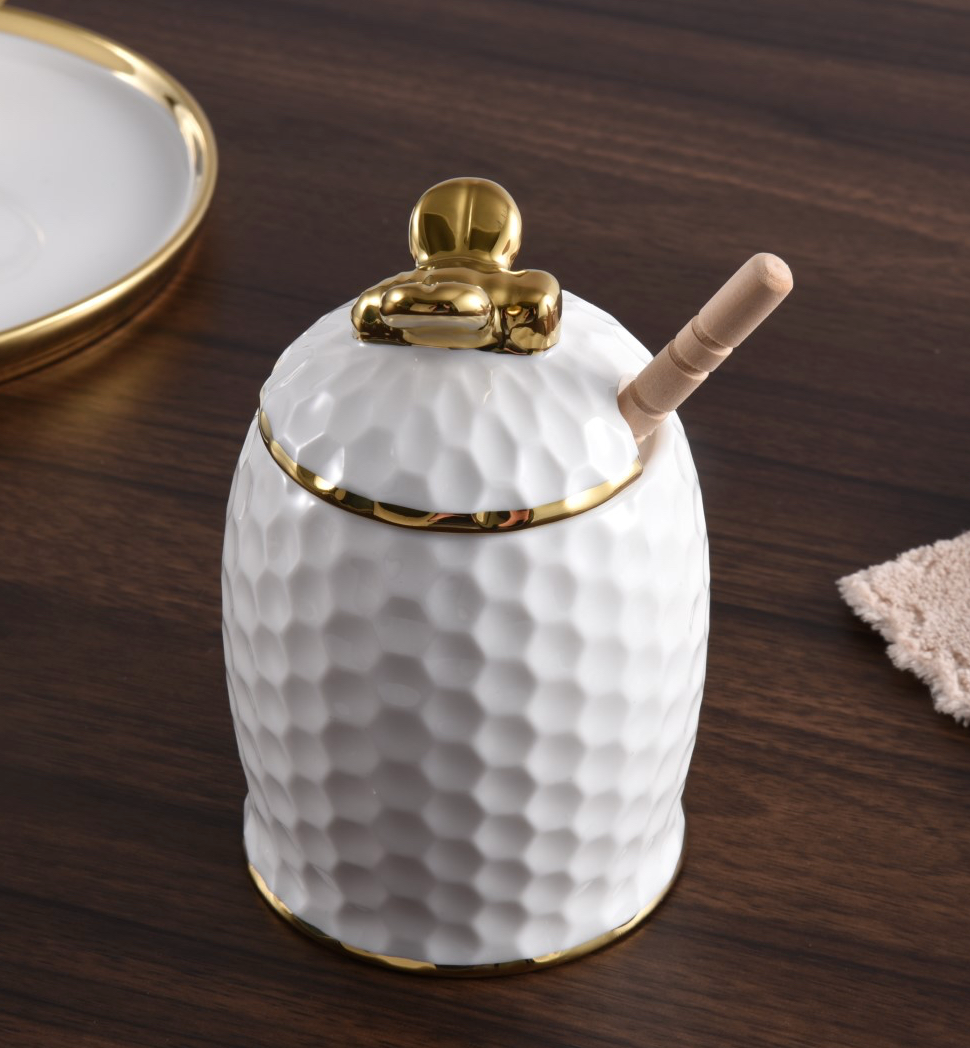 The PAMPA BAY Honey Jar Set features a white porcelain honey jar adorned with a textured, honeycomb-inspired surface and detailed gold accents. A gold bee-shaped handle tops the lid, while a wooden dipper rests inside. Presented on a dark wooden table alongside a plate and napkin, this elegant set makes an exquisite hostess gift for any occasion.
