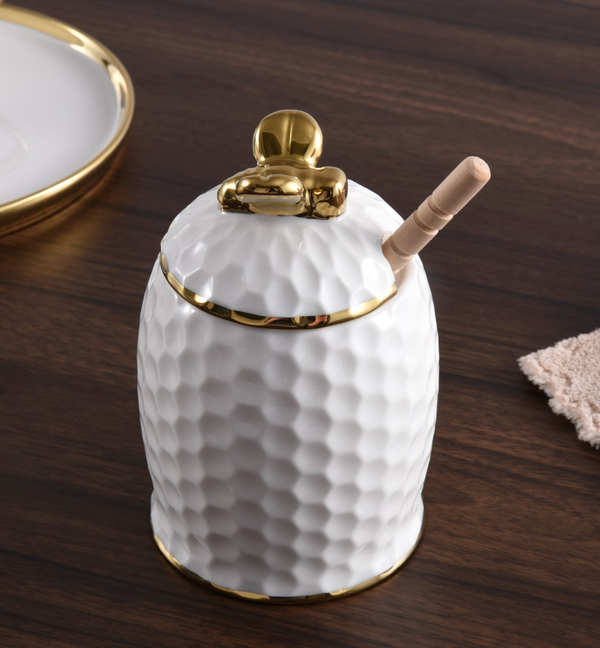The PAMPA BAY Honey Jar Set features a white porcelain honey jar adorned with a textured, honeycomb-inspired surface and detailed gold accents. A gold bee-shaped handle tops the lid, while a wooden dipper rests inside. Presented on a dark wooden table alongside a plate and napkin, this elegant set makes an exquisite hostess gift for any occasion.