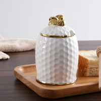 A luxurious hostess gift for any occasion, the PAMPA BAY Honey Jar Set features a white, textured porcelain jar with a gold lid, displayed on a wooden tray beside sliced bread and a honey dipper, with a soft beige cloth in the background.