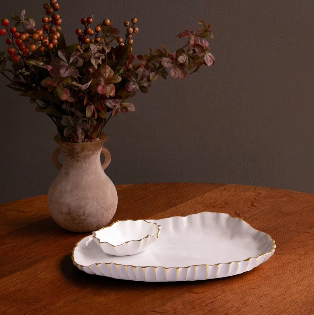 A ceramic vase with red and green foliage is on a wooden table beside two white, scalloped plates with gold trim and the Beatriz Ball Thanni Mystic Oval Platter w/ Bowl White & Gold - Large, set against a plain dark wall.