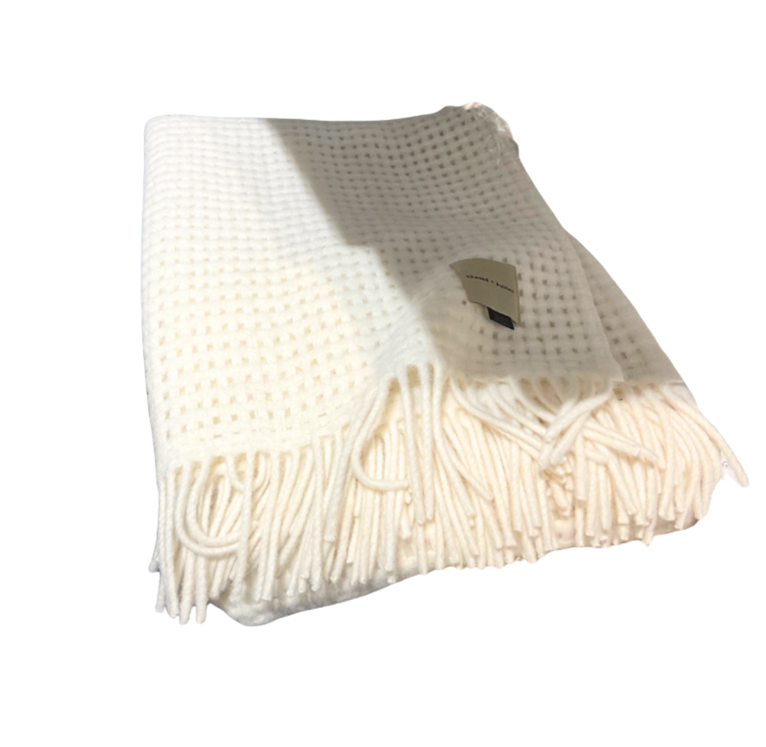 The Thread & Butter Waffle Alpaca Throw - Ivory is a cream-colored, sustainable alpaca blanket with a waffle texture, fringed ends, and a small tag, shown folded on a white background.