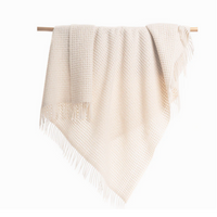 The Thread & Butter Waffle Alpaca Throw - Ivory features a cream hue, textured waffle pattern, and fringe ends, draped over a wooden rod against a white backdrop. This sustainable blanket brings warmth and elegance to any room.