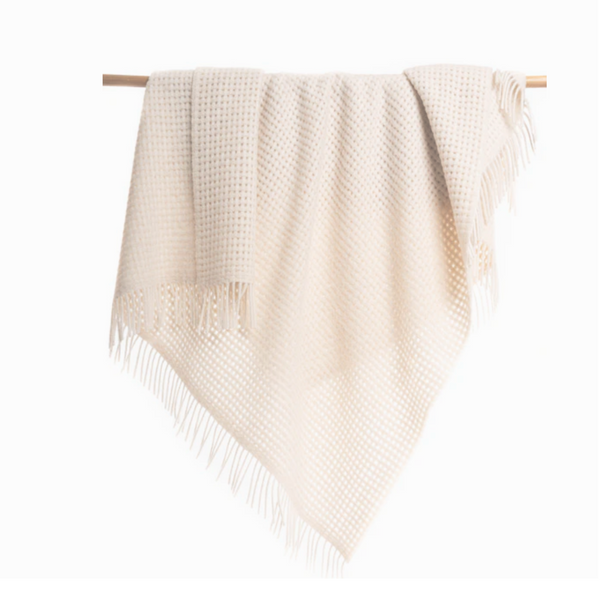 The Thread & Butter Waffle Alpaca Throw - Ivory features a cream hue, textured waffle pattern, and fringe ends, draped over a wooden rod against a white backdrop. This sustainable blanket brings warmth and elegance to any room.
