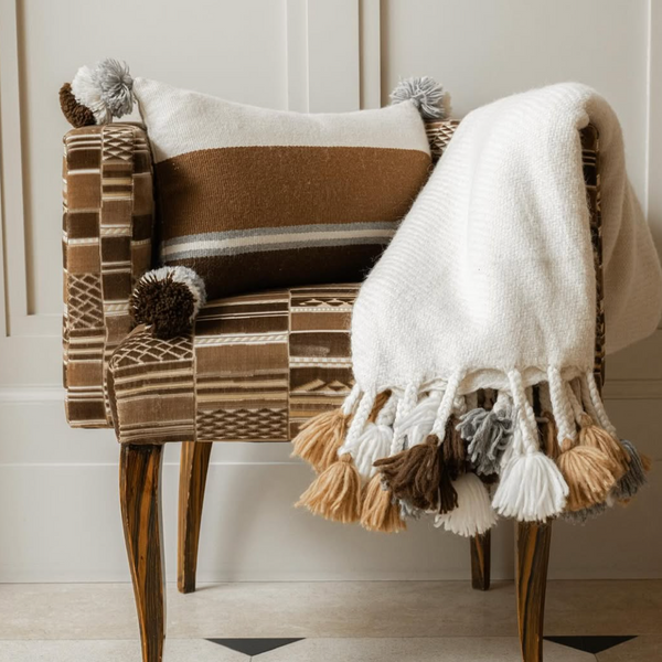 A patterned brown armchair with wooden legs features a rectangular brown and white pillow and the Cusco Throw by Threads Soft Goods, an alpaca blanket adorned with large beige, brown, and gray tassels.