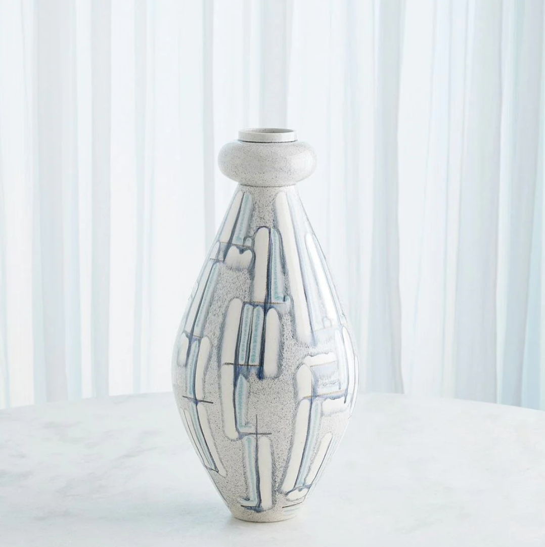 The Spindel Vase Grey Blue by Global Views/Surya, a tall white Portuguese ceramic piece with blue and gray linear patterns, stands on a round white surface before sheer vertical white curtains.