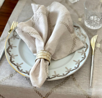 A close-up of a formal table features Bodrum’s Regent Napkin Tan Set of 4, elegantly displayed with a napkin ring, gold-handled flatware, a decorative plate, clear glassware, and beautifully embroidered placemats.
