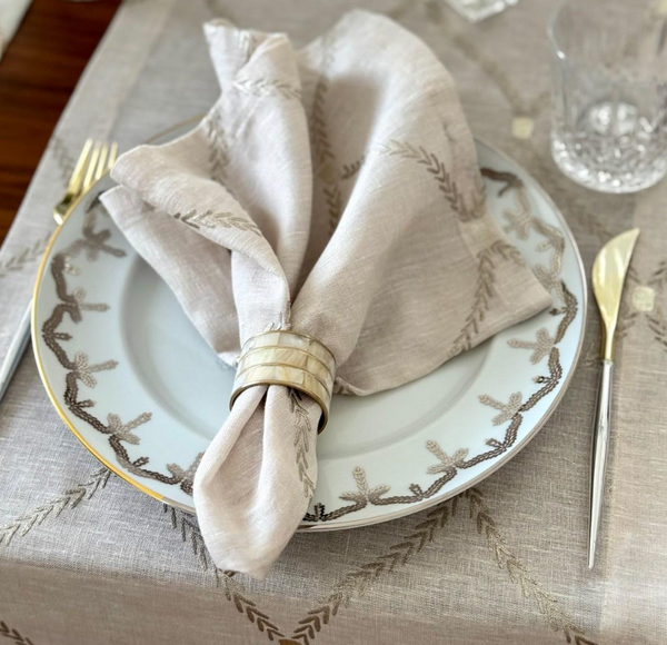 A close-up of a formal table features Bodrum’s Regent Napkin Tan Set of 4, elegantly displayed with a napkin ring, gold-handled flatware, a decorative plate, clear glassware, and beautifully embroidered placemats.