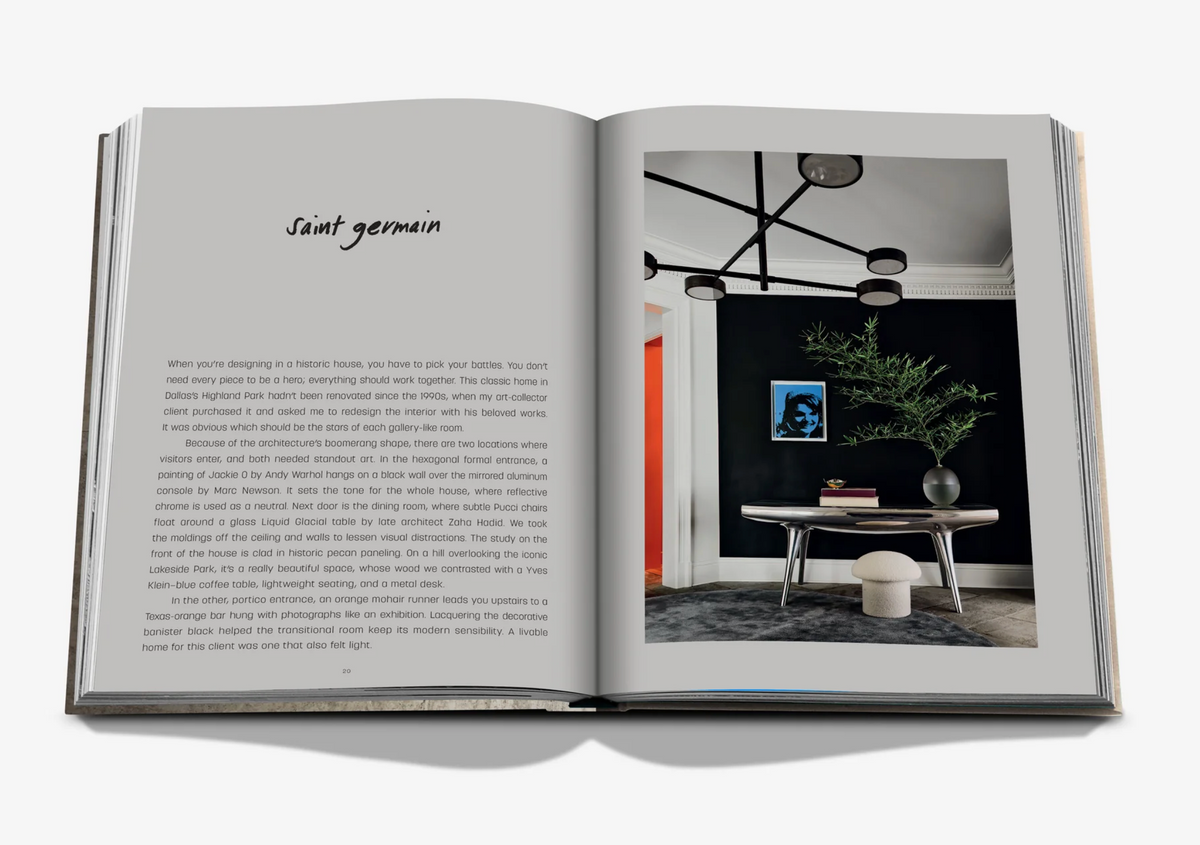An open book titled "Relaxed Luxury" by Assouline displays the Saint Germain room description on the left and a modern interior by Chad Dorsey on the right, featuring a table, plant, artwork, round stool, and black geometric chandelier.