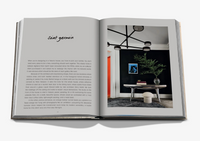 An open book titled "Relaxed Luxury" by Assouline displays the Saint Germain room description on the left and a modern interior by Chad Dorsey on the right, featuring a table, plant, artwork, round stool, and black geometric chandelier.