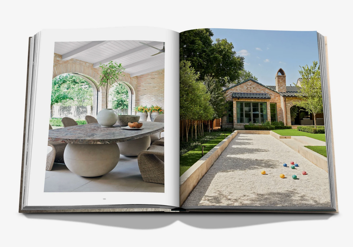 The Assouline book "Relaxed Luxury" opens to a sunlit dining area with arched windows and a stone table on one page, and a landscaped garden with a modern brick house and gravel yard on the other.