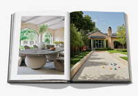 The Assouline book "Relaxed Luxury" opens to a sunlit dining area with arched windows and a stone table on one page, and a landscaped garden with a modern brick house and gravel yard on the other.