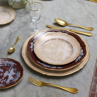 A formal marble table is set with Ginori 1735’s Oriente Italiano Castagna tableware in beige and burgundy with white patterns, gold-colored cutlery, and a clear stemmed glass. Italian craftsmanship is evident in every detail.